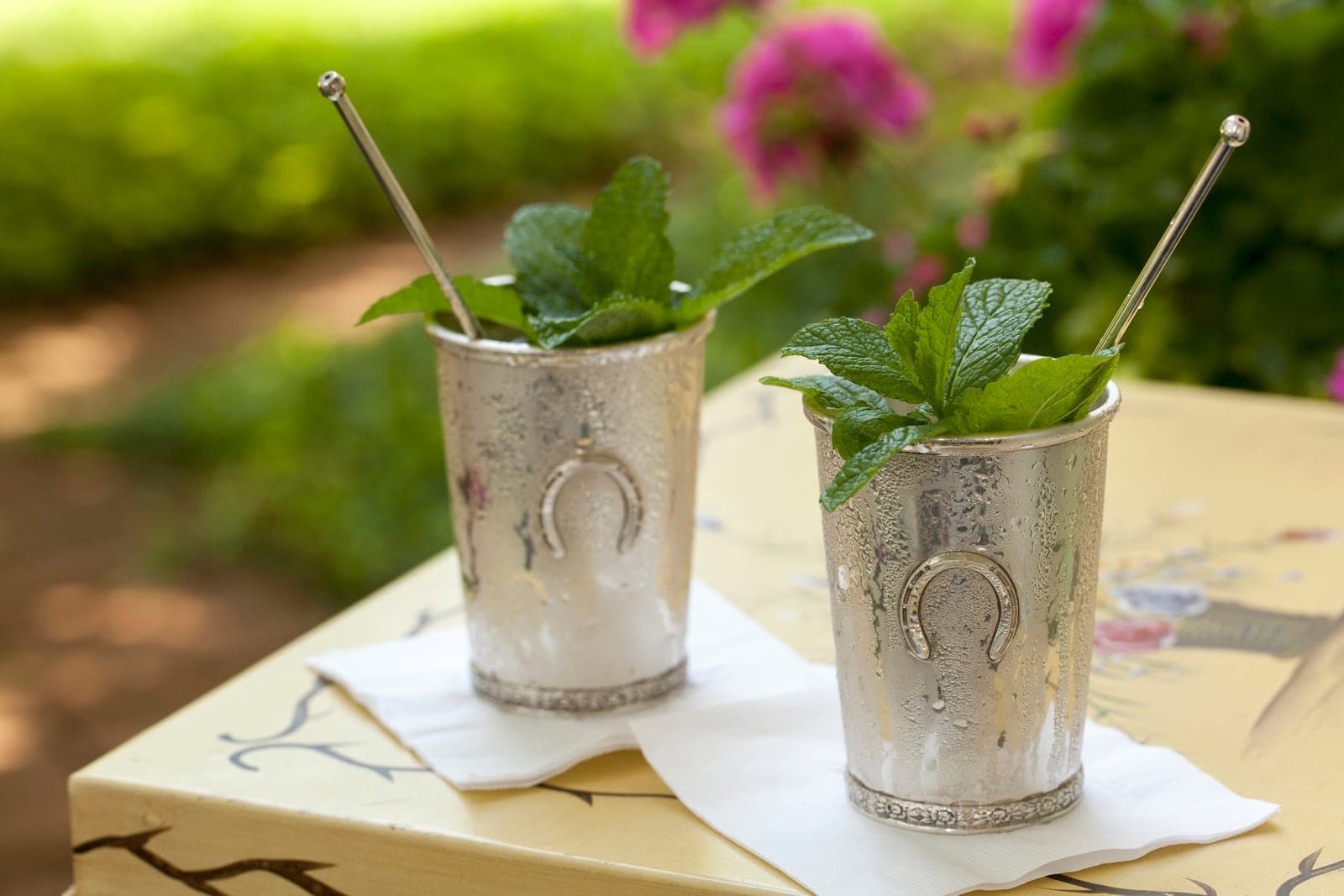 Two silver mint juleps with horseshoe medallions, served in the garden.